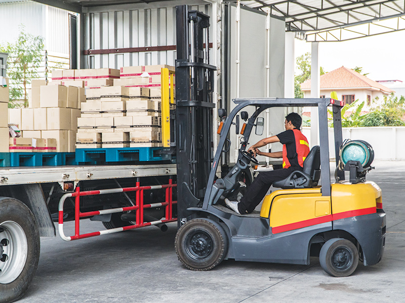 Warehouse worker loading a flat bed truck using a propane powered forklift with propane tank visible on back of forklift