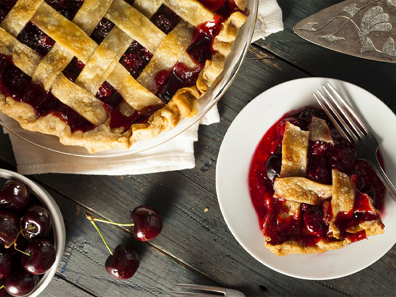 a slice of cherry pie on a plate with a fork next to a whole lattice-top cherry pie