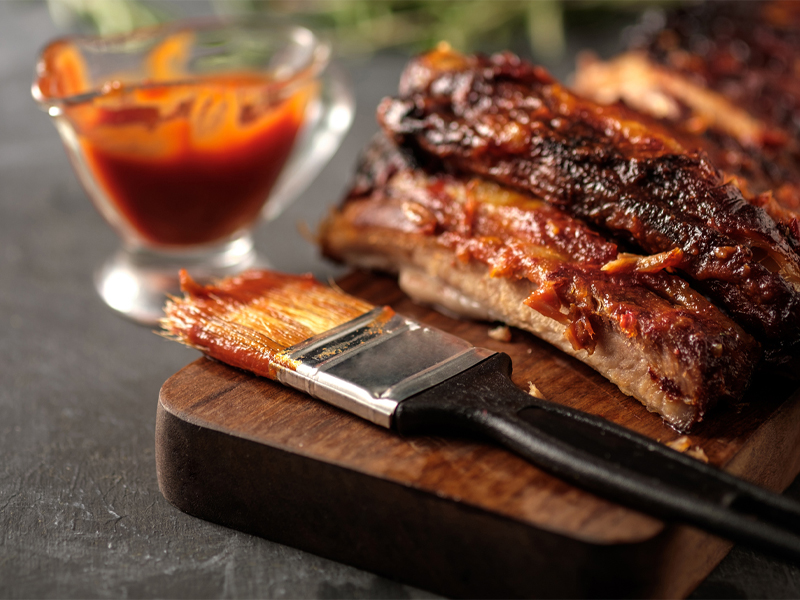 Chocolate Chipotle BBQ sauce in a clear dish next to glazed ribs and a pastry brush