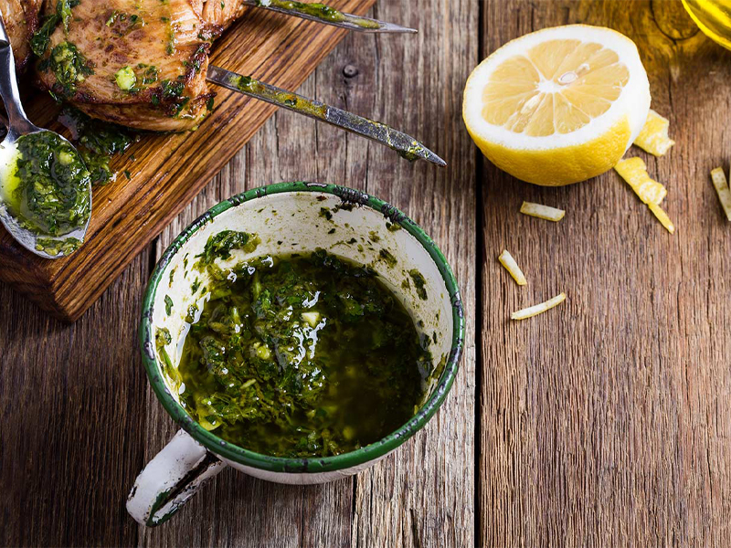 Cup of chimichurri on a board with a fresh lemon and grilled chicken in the background