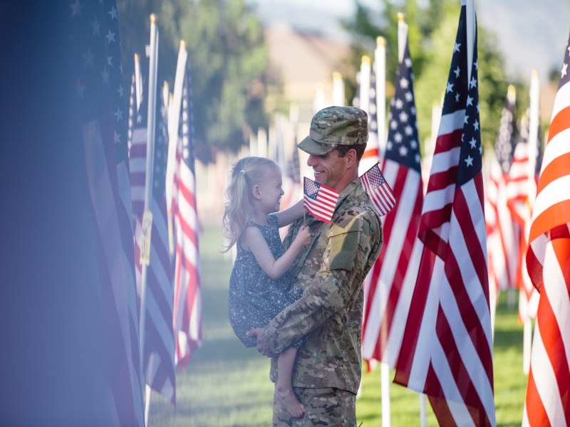 Man dressed in uniform holding a girl in a field of American Flags