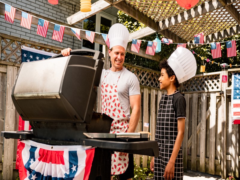 Man and boy in hats and aprons cooking out on a grill with patriotic decor in the background