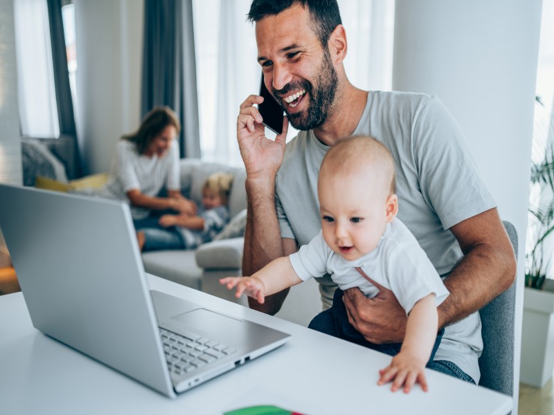Man on the phone by his laptop, holding a baby